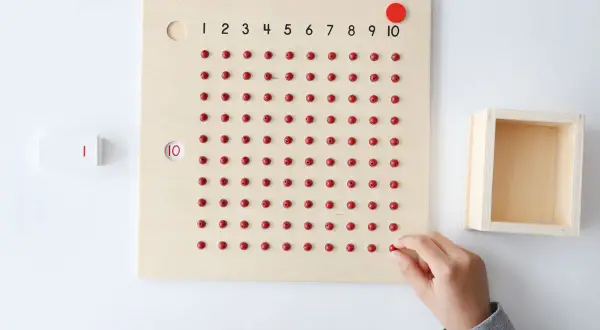 Tabla de fichas de multiplicaciones con cuentas rojas y números. Una mano coloca una cuenta, aprendiendo las tablas.