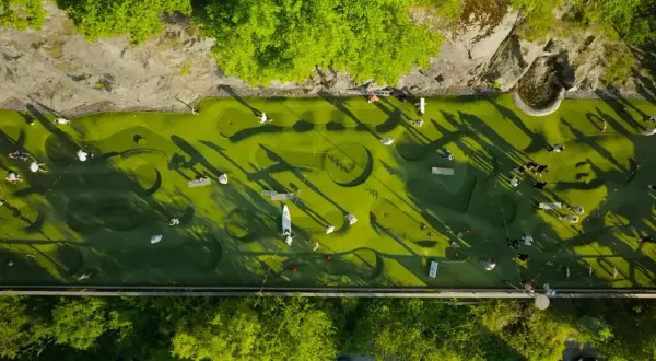 Folk spelar minigolf i Nacka Strand. En grön bana med hinder och skuggor från träden.