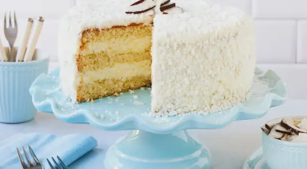 Eine köstliche Kokos Torte, bedeckt mit Kokosraspeln und dekoriert mit Kokoschips, wartet auf dem hellblauen Tortenständer.