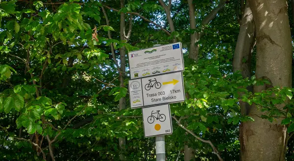 Znaki szlaku rowerowego wskazują trasę 003 do Starego Bielska, 17 km. W tle zielone drzewa.