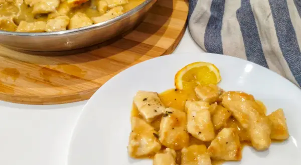 Petto di pollo al limone cremoso servito su un piatto bianco, con una fetta di limone e una padella piena dello stesso piatto sullo sfondo.