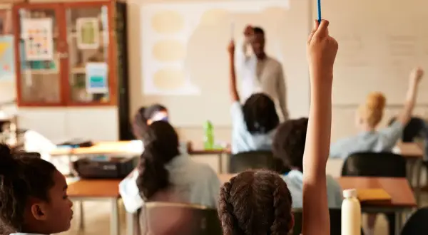 Estudiantes levantan la mano en clase. El profesor proyecta información, quizás sobre el calendario escolar 2026-27.