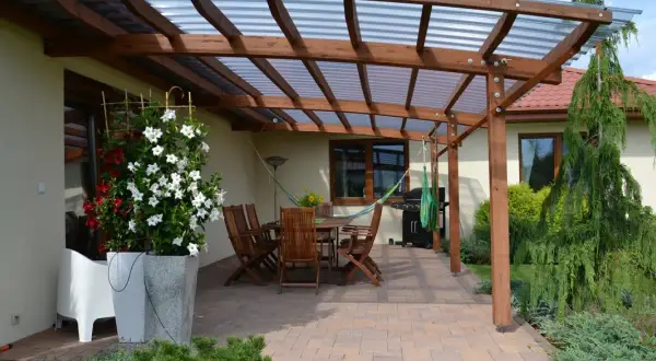 Drewniana pergola z przezroczystym dachem nad tarasem, gdzie stoi stół z krzesłami, grill i hamak. Idealne miejsce, co na taras.