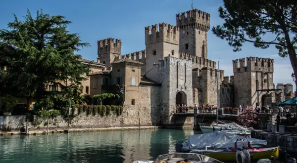 Il castello di Moniga si affaccia sull'acqua, con torri merlate e un ponte pedonale affollato. Barche ormeggiate completano la scena.