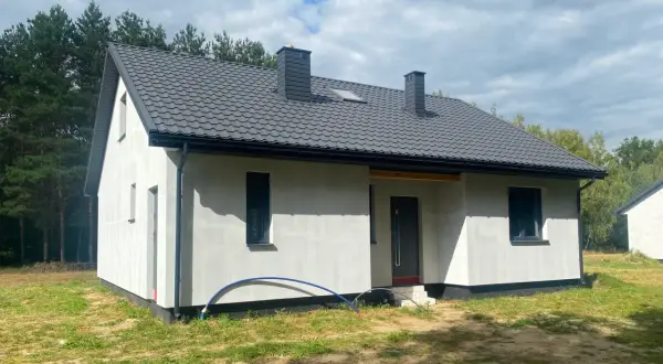 Nowy, biały dom szkieletowy z szarym dachem i dwoma kominami, otoczony zielenią.