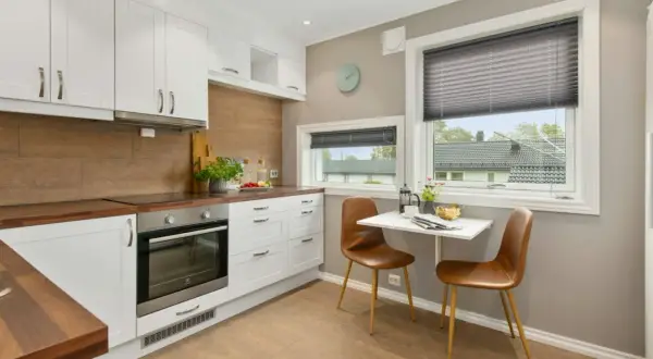 Moderna cocina equipada con gabinetes blancos, encimeras de madera y un horno. Una pequeña mesa con sillas marrones está junto a la ventana.