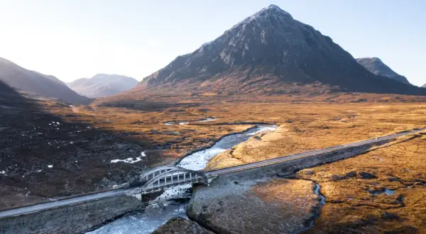 Majestätische Berge der Schottland Highlands, ein Fluss schlängelt sich durch das goldene Gras, überquert von einer Brücke.