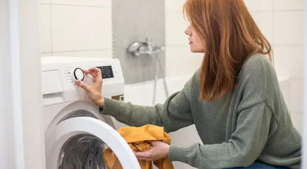 Frau stellt Waschmaschine ein, um den Wasserverbrauch zu optimieren. Sie hält ein gelbes Kleidungsstück in der Hand.