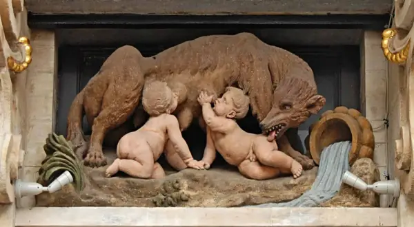 Rzeźba przedstawia wilczycę karmiącą Romulusa i Remusa, symbolizującą legendę o założeniu Rzymu. To klasyczny przykład motywu salut rzymski.