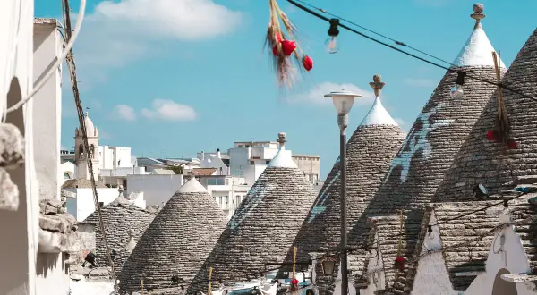 Scorci di Alberobello con i caratteristici trulli, alcuni decorati con simboli bianchi, sotto un cielo azzurro. Una scalinata cuori conduce tra le abitazioni uniche.