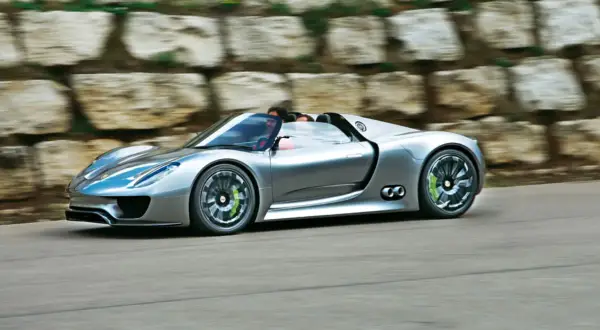 Srebrny Porsche 918 Spyder pędzi po drodze, z kierowcą za kierownicą.