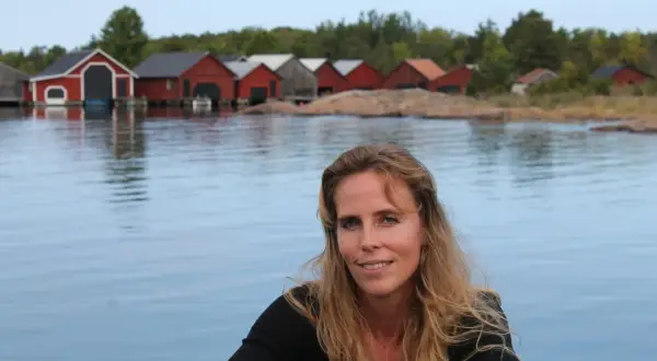 Woman with long blonde hair smiling, with red boathouses on a rocky shore in the background.
