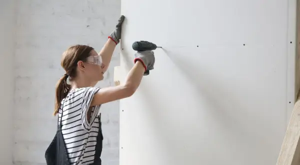 Mujer con gafas de seguridad y guantes instala placas de yeso, creando un trasdosado.