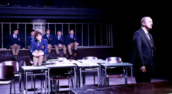Scena z "Stowarzyszenia Umarłych Poetów" w teatrze. Uczniowie w mundurkach siedzą na ławce, jeden z nich na pierwszym planie. Nauczyciel stoi po prawej.
