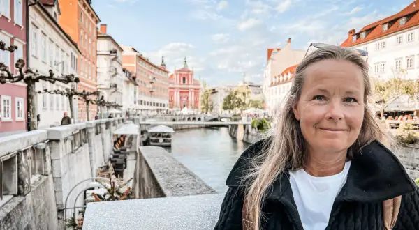 Kvinna ler vid floden i Ljubljana, Sloveniens huvudstad, med färgglada byggnader och en kyrka i bakgrunden.