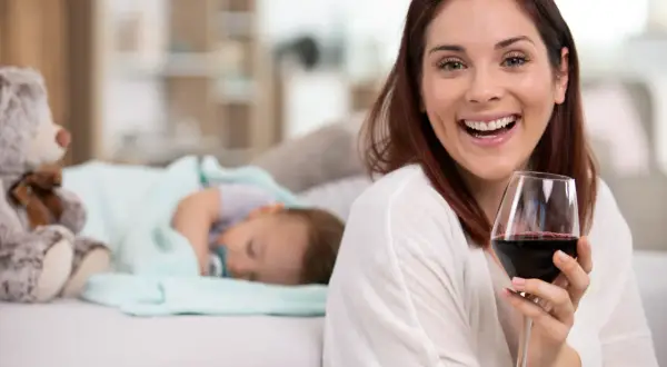 Uśmiechnięta kobieta z kieliszkiem czerwonego wina, podczas gdy dziecko śpi obok. Temat: karmienie piersią a alkohol.