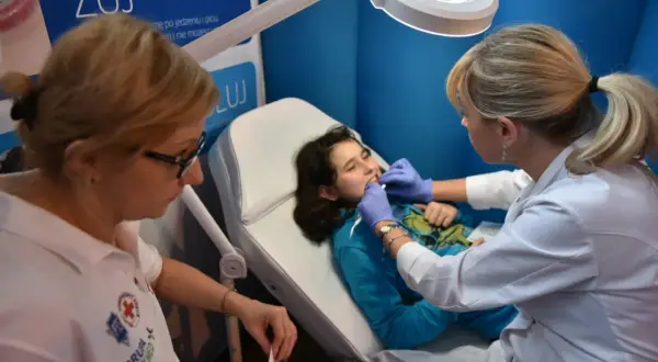 Dziecko na fotelu stomatologicznym, badane przez lekarkę. Na plakacie w tle widnieje napis "ŻUJ" i logo Luxmed. Sprawdź luxmed stomatologia cennik.
