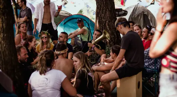 Grupa ludzi relaksuje się na festiwalu Pannonica, jeden z nich gra na puzonie. W tle widać namioty.