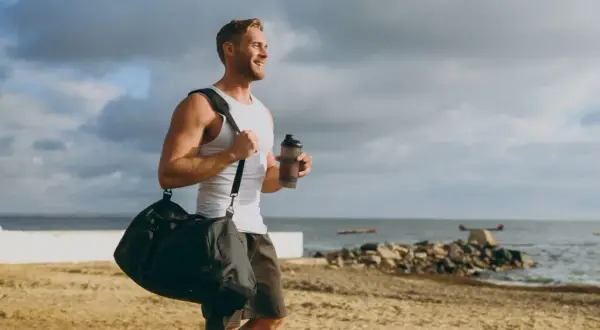 Uśmiechnięty mężczyzna z torbą sportową i bidonem na plaży. Zastanawia się, glutamina co to, czy pomoże mu w regeneracji po treningu.