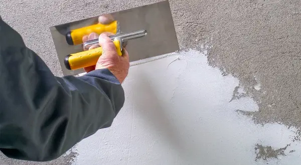 Mano con spatola gialla applica pittura rasante su muro grezzo, creando una superficie liscia e uniforme.