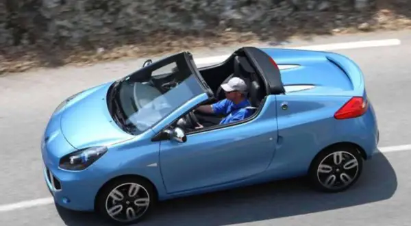 Niebieski kabriolet Renault Wind z otwartym dachem, kierowca w czapce i niebieskiej koszulce jedzie po krętej drodze.