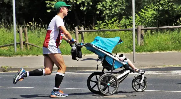 Mężczyzna w zielonej czapce i sportowym stroju bierze udział w biegu z wózkiem. Dziecko w wózku ma na sobie białe skarpetki i sandałki.