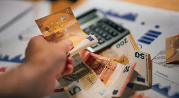 Dłoń trzyma banknot 50 euro nad stosem innych banknotów i kalkulatorem. To symbol prowizji i finansów.