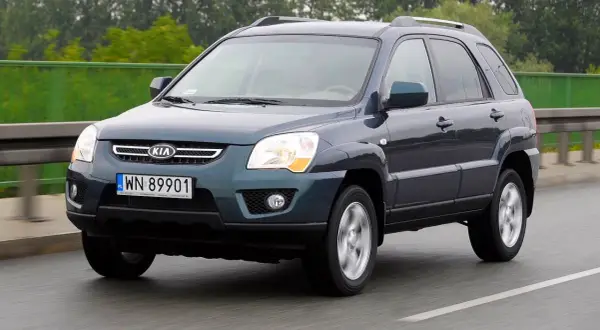 Ciemnoszary Kia Sportage 2008 na drodze, z zielonym ogrodzeniem w tle.