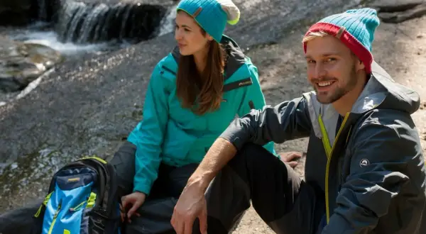 Para, co to jest trekking? Para w czapkach i kurtkach siedzi nad strumieniem, obok plecak.