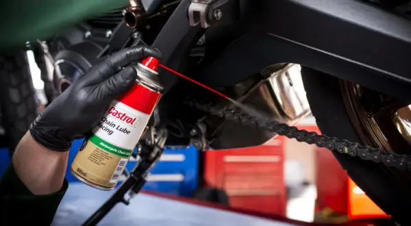 Ręka w czarnej rękawiczce rozpyla smar Castrol na łańcuch motocykla.