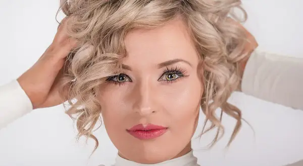 Kobieta z kręconymi blond włosami, jej spojrzenie jest nonszalanckie, a dłonie unoszą się ku głowie.