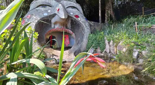 Scultura di una testa gigante con bocca spalancata nel giardino botanico Gardone Riviera, con piante rigogliose e uno stagno.