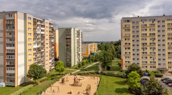 Bloki mieszkalne z placem zabaw na pierwszym planie, pod pochmurnym niebem.