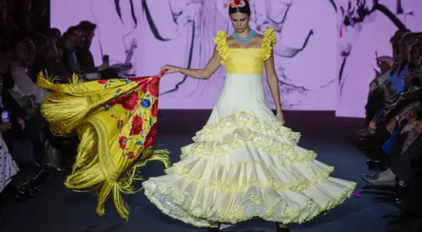 Modelo desfila en la pasarela We Love Flamenco con un vestido blanco de lunares amarillos y volantes, portando un mantón amarillo con flores rojas.
