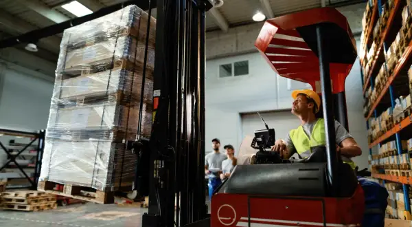 Operator wózka widłowego przygotowuje transport drobnicowy. Na paletach leżą paczki owinięte folią.