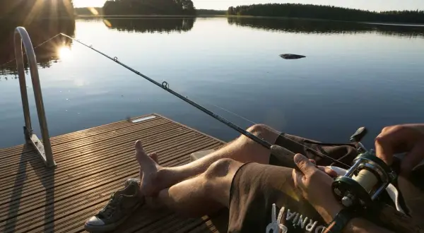 Słońce zachodzi nad spokojnym jeziorem. Wędkarz siedzi na pomoście, trzymając wędkę, gotowy na złapanie ryby.