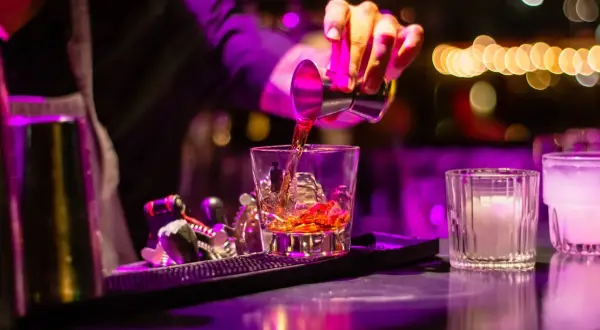 Barman nalewa bursztynowy cordial drink do szklanki z lodem. W tle kolorowe światła.