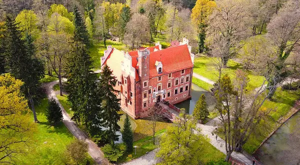 Zamek na wodzie w Wojnowicach otoczony wiosenną zielenią i drzewami. Czerwona bryła zamku z charakterystycznym dachem odbija się w spokojnej tafli wody.
