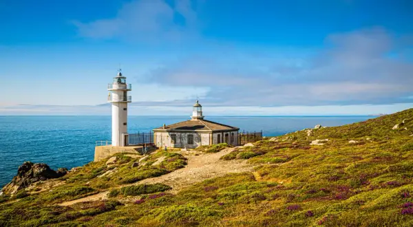 Faro blanco y edificio antiguo en Cabo Touriñán, con mar azul y cielo nublado.