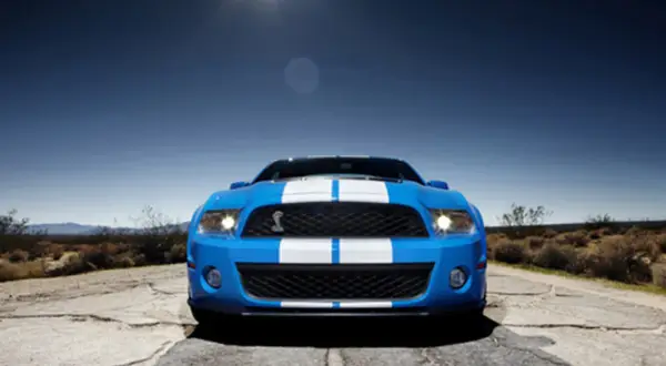 Niebieski Shelby GT500 z białymi pasami na masce i dachu, zaparkowany na pustynnej drodze pod błękitnym niebem.