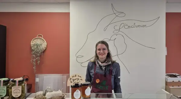 Une vendeuse souriante derrière un comptoir, avec des cornichons, du miel et du fromage de chèvre, peut-être du fromage Saint-Aubin, en vente.