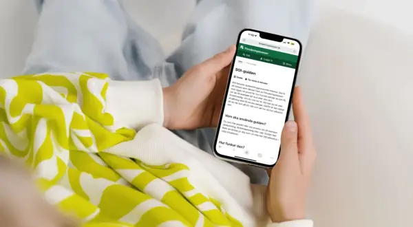 Person håller upp en mobiltelefon som visar Försäkringskassans SGI-guide. Guiden förklarar vad SGI är och hur den fungerar för enskild firma.