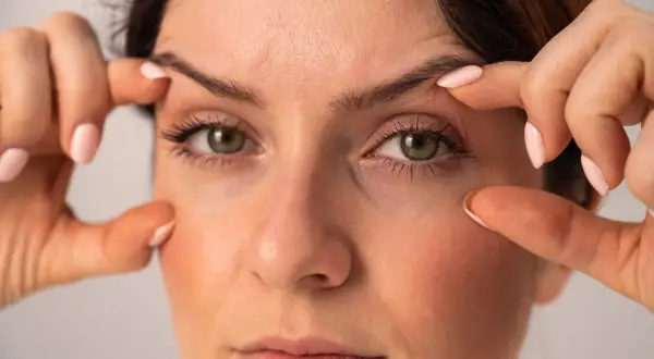 Une femme examine son visage, ses doigts effleurant sa peau. Elle semble préoccupée car j'ai des cernes noires sous les yeux.