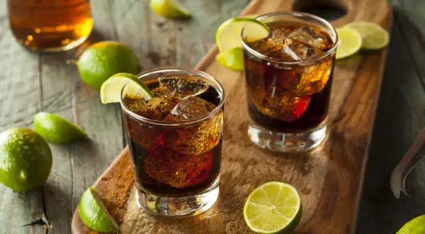Dwa drinki z rumem i colą, ozdobione ćwiartkami limonki, leżą na drewnianej desce. Idealne z czym pić rum!