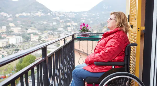 Donna in carrozzina su un balcone, con vista su un paesaggio collinare. Un'immagine che evoca la necessità di un piano nazionale non autosufficienza.