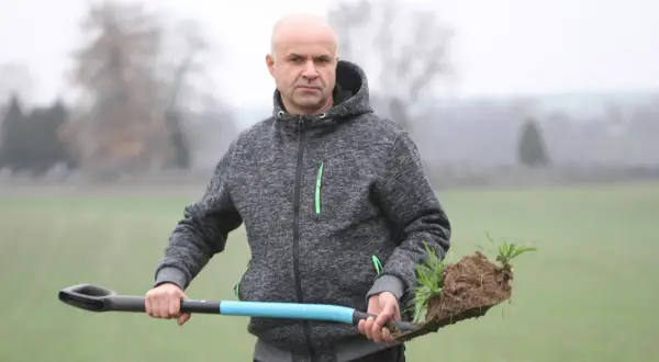 Mężczyzna w szarej bluzie z kapturem trzyma łopatę z bryłą ziemi i młodymi roślinami. Może to być sadzonka jęczmienia.