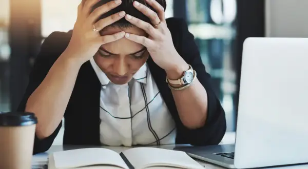Kvinna med händerna i håret, blicken sänkt över en öppen bok. Känslan av stress på jobbet är påtaglig. En kaffekopp och laptop finns i närheten.