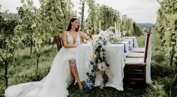 Braut in weißem Kleid sitzt am Tisch im Weinberg. Eine romantische Kulisse für eine weingut hochzeit.