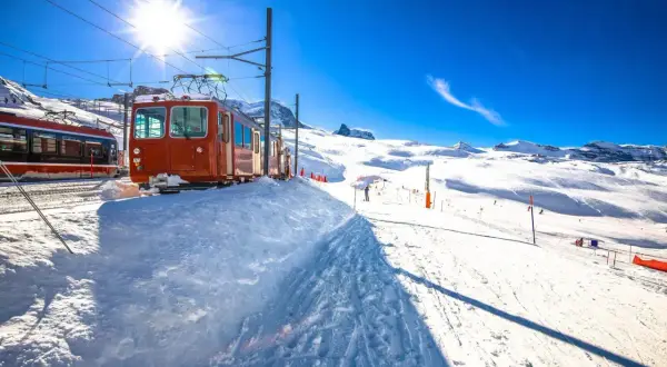 Bernina Express-tåget står parkerat i snön, redo att ta passagerare genom ett snöigt alpint landskap under en klarblå himmel.
