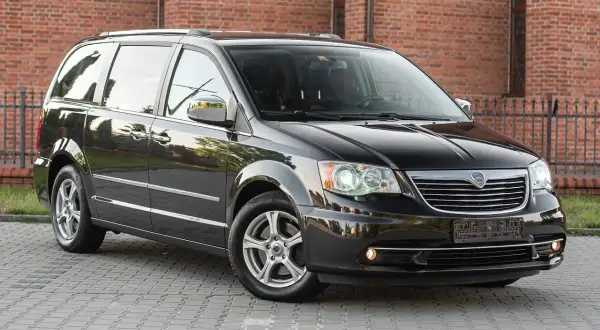Czarny Lancia Voyager stoi na brukowanym podjeździe przed ceglanym budynkiem.